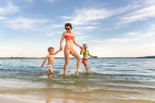 Young adult attractive slim sporty mother enjoy having fun running water by lake or sea sand beach with two cute little siblings against blue sky on summer day. Summertime family vacation concept