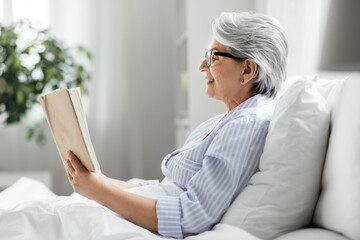 technology, old age and people concept - happy smiling senior woman in glasses reading book in bed...