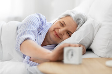 old age and people concept - happy smiling senior woman with alarm clock lying in bed at home...