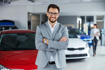 Smiling car seller standing in car salon with arms crossed and waiting for customers to come on...