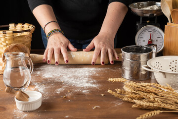 View of woman's hands with rolling pin on wooden table with flour, kitchen objects, wheat, weight, black background, horizontal