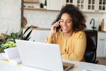 Delighted happy curly African-American young woman talking on the smartphone and using laptop at home, student girl studying at home, has phone conversation, takes a break and chatting with a friend
