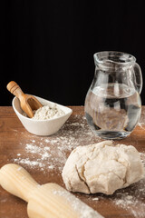 Top view of bowl with flour, pizza dough, jug of water and rolling pin, with selective focus, on wooden table with flour, black background, vertical