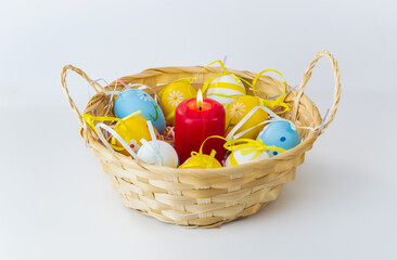 Fototapeta na wymiar Easter composition: a straw basket, colored Easter eggs lying in it, a burning red candle on a light background.