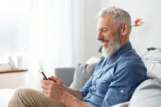Grey-haired middle aged hipster man in casual shirt using smartphone sitting on the sofa at home, mature guy enjoys online chatting, spends time in social networks, looks at the phone and smiles