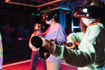 african american teenage girl in vr headset and safety mask gaming near blurred friends