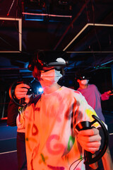 Fototapeta na wymiar teenage boy in medical mask gaming near blurred friends in vr play zone