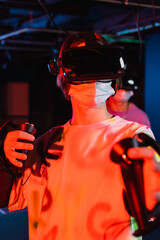 teenage gamer in medical mask and vr headset near friend on blurred background