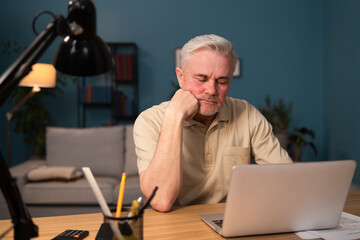 A tired, broken, resigned, preoccupied elderly man sits in front of a laptop, computer and covers...
