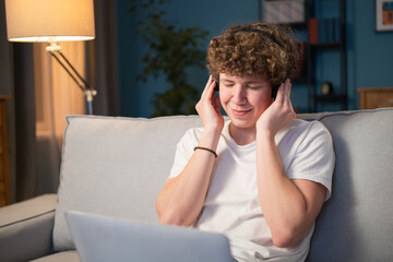 Fototapeta premium A young boy sits on a couch in the living room with a laptop on his lap and wireless headphones on his ears on which he holds his hands. The teenager is listening to music with his eyes closed.