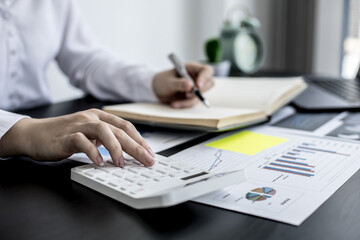 An accounting businesswoman pressing the white calculator and taking notes in her notebook, she was examining the company's financial accounts from the documents provided by the finance department.