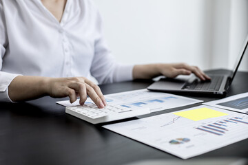 A woman uses a laptop and presses a white calculator, she is a company auditor, she checks documents from the laptop and calculates the numbers for accuracy. Financial audit concept