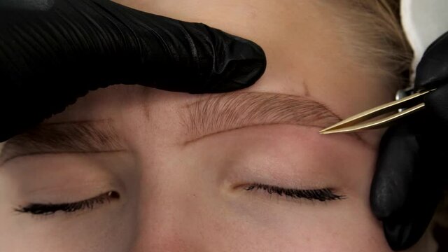 Gloved master's hands pluck model's eyebrows with tweezers close-up. Eyebrow lamination and styling. Shape of the eyebrows is drawn with a pencil on the face of a beautiful young woman. 