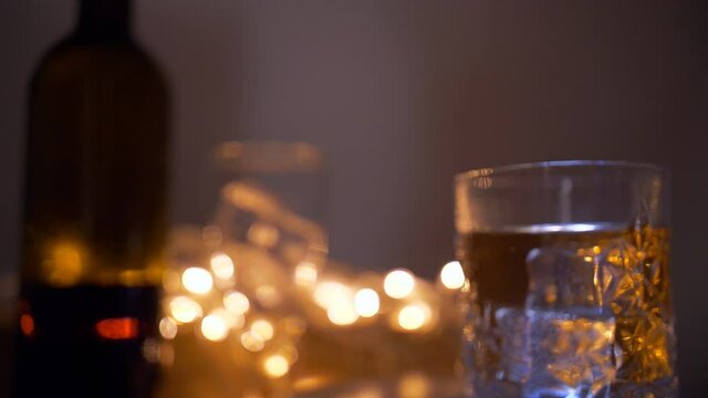 Racking shot of a glass of whiskey filled with ice cubes ready to drink. Beautiful view of crystal glass of whiskey/hard drink with an alcohol bottle and decorative lights in the background
