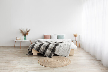 Bed with pillows and blanket, round carpet, tables with dry plants in vases on wooden floor
