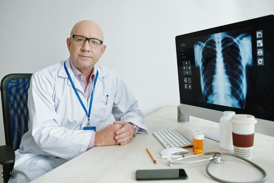 Portrait of serious mature bald doctor in lab coat sitting at desk and working with online x-ray image on computer