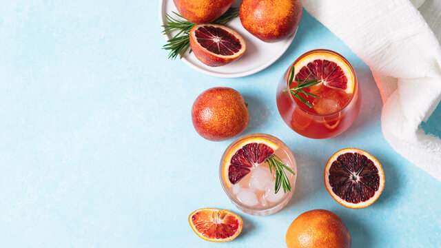 Blood orange cocktails with fresh fruits on blue table background. Summer cocktails, lemonade, refreshing drinks, low alcohol mocktail, party concept. Banner, flat lay, top view, copy space.