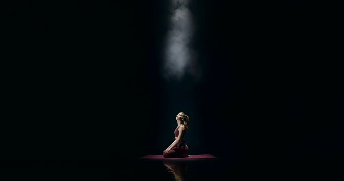 A sportive woman sits on a mat on stage and stretches her thigh muscles. It is dark around, but you can see her silhouette, because a spotlight is shining on the girl. She relaxes and meditates