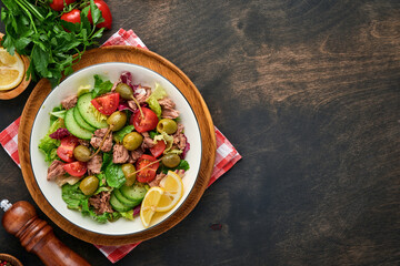 Tuna salad with fresh vegetables, olives, capers and lemon served in bowl on old wooden background. Top view with copy space.