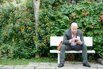 Fototapeta premium Serious bald mature businessman in formal suit sitting with gadget in park and drinking coffee while having break