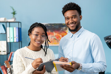 A woman with glasses and a man with stubble, happy to be working for the company are smiling at camera, holding documentation, notepads and pens in hands. Office in the background, filling cabinet