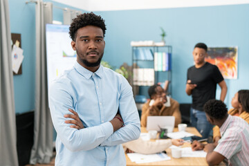 Portrait of a serious dark-skinned man wearing a shirt with his arms crossed over his chest, the guy has dark hair and stubble, a business meeting of a group of employees is going on in the background