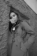 gorgeous asian attractive model wearing trench coat standing near wall shot from lower point. outdoors black and white portrait. young beautiful sensual female looking at camera, holding glasses