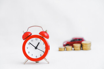 close up of analog red clock with blurred miniature red car on stack of golden coins on grunge light grey background, new car, lower price, sale automobile, reduced price concept