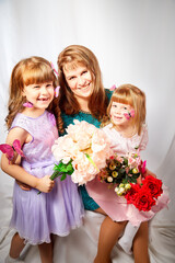 Cute daughters and mother with flowers in the studio on a white background. Young girls and woman posing indoors. Family during photo shoot in summer or spring time