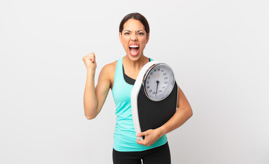 young hispanic woman shouting aggressively with an angry expression and holding a scale