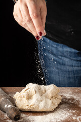 Close-up of woman's hand pouring flour on pizza dough, on wooden table with flour and rolling pin, black background, vertical