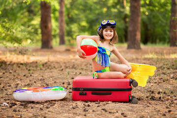Enthusiastic little girl in swimsuit, fins and swimming mask with circle and swimming ball packed suitcase, and ready to fly for summer vacation trip