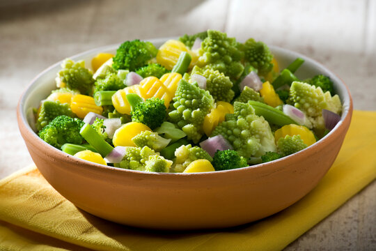 Variety of steamed healthy vegetables in a bowl on table. Mixture of broccoli, romanesco, yellow carrots, red onions.