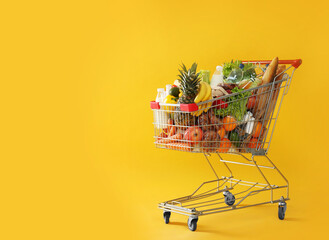 Shopping cart full of groceries on yellow background. Space for text - obrazy, fototapety, plakaty