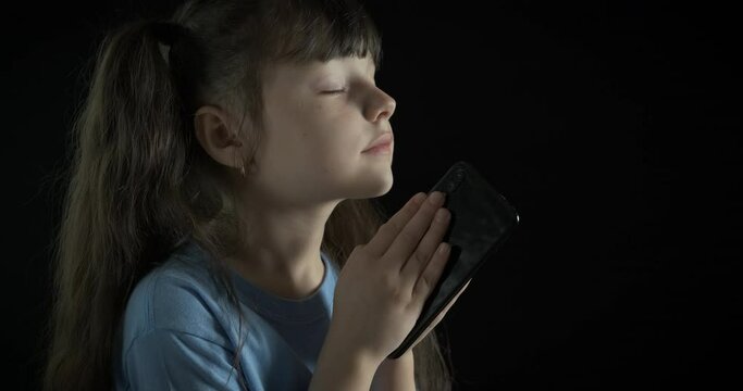 Pray with a telephone. A child with a telephone in her hands pray on her gadget. A concept of believing in smart phones.