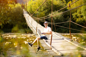 Concentrated young businessman in eyewear sit on bridge across mountains river with laptop. Web surfing information in internet or working distantly online. Communication remotely with client or study