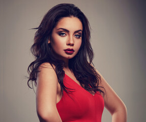 Fototapeta premium Beautiful makeup woman in red dress with long black curly volume hair style, burgundy lipstick with vamp look on grey background. Closeup