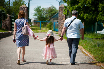 Naklejka na ściany i meble family walking in the park. mom dad and little girl walking holding hands. back view. daughter in a pink dress. friendly loving family concept, love. outdoor, walk in the fresh air, threesome