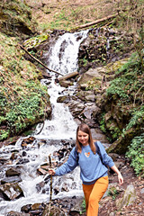 Naklejka premium Young blond woman with stick in her hand going down stones along small waterfall in the mountain forest, mountain river, active lifestyle, digital detox, hiking alone