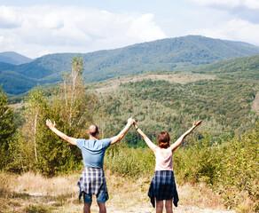 Back view of two young people holding her hands up standing together at the top of the mountain. Sunny and bright autumn day. Hiking concept. Concept of freedom.