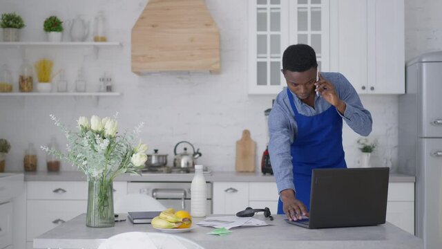 Serious confident African American man in blue apron talking on the phone standing in kitchen. Portrait of concentrated smart young startuper discussing business idea from home office