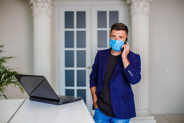 A young man in a medical mask and a blue jacket talking on the phone and working on a laptop in a bright room. internet office