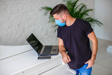 A young man in a medical mask and a black T-shirt, working on a laptop in a bright room. internet office