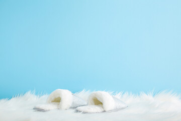 Beautiful soft warm female slippers on white fluffy fur carpet at light blue wall background. Pastel color. Closeup. Comfortable home shoes. Empty place for text. Front view. - obrazy, fototapety, plakaty