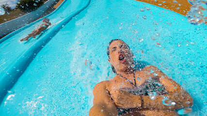 Selfie 30s woman sliding a water slide very fast, splashing into the pool with water drops on the...