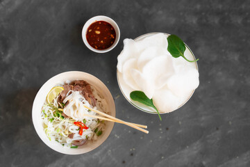 Pho bo, a traditional Vietnamese noodle and beef soup and a traditional snack of rice chips and sesame sauce on a gray background.