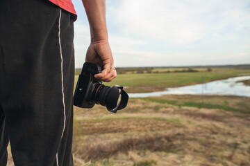 Fototapeta premium A male amateur photographer with a camera stands on a hill near a green meadow at sunset. Photorapher landscaper