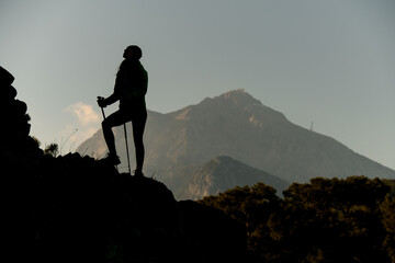 view of dark silhouette of woman with trekking poles against background of sky and mountains