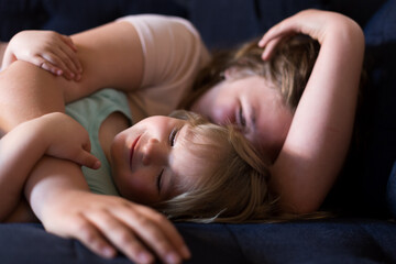 Horizontal selective focus view of cute blond dishevelled toddler girl cuddling lying down on couch in the arms of older sister in soft focus background