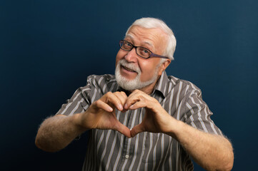 Fototapeta na wymiar elderly gray-haired man in glasses and black shirt shows hands heart symbol-love, emotions in frame, waist portrait, blue background, studio
